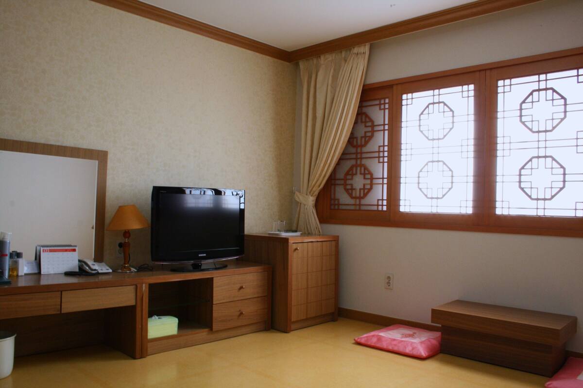 A hotel room with a wooden desk, a television, a small cabinet, a window with decorative glass, and a pink cushion.