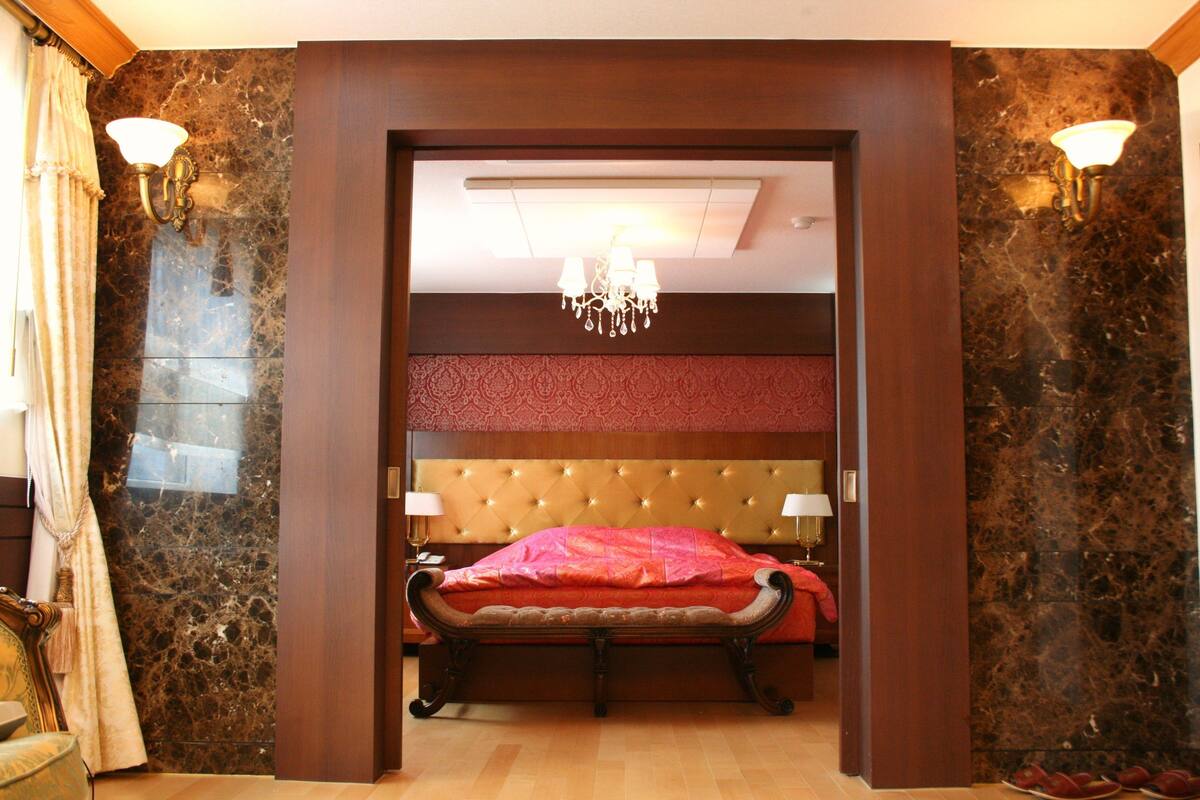 A bedroom with a tufted headboard, a bed with a red bedspread, two wall-mounted lamps, and a chandelier.