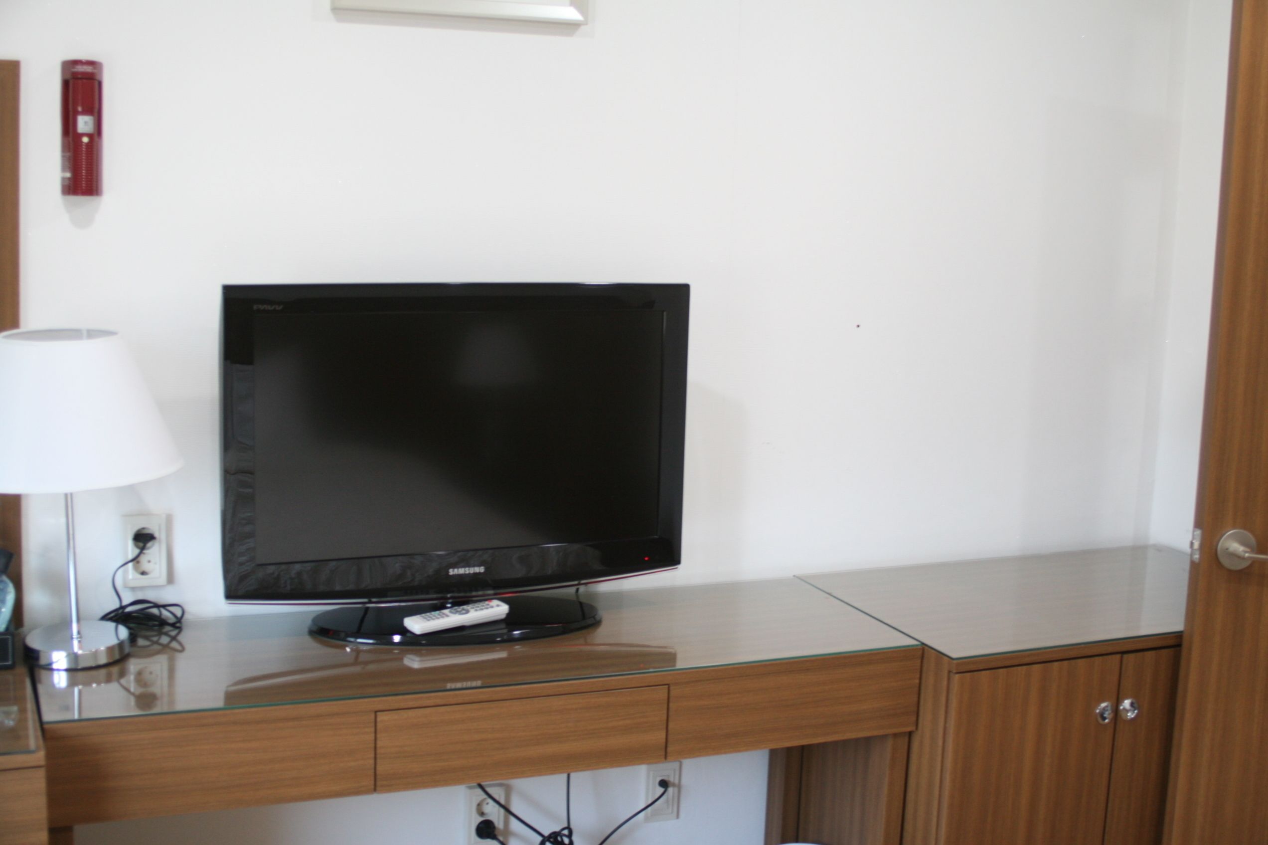 Una habitación de hotel moderna con una televisión de pantalla plana sobre un escritorio de vidrio, una mesita de noche con lámpara y un armario de madera.