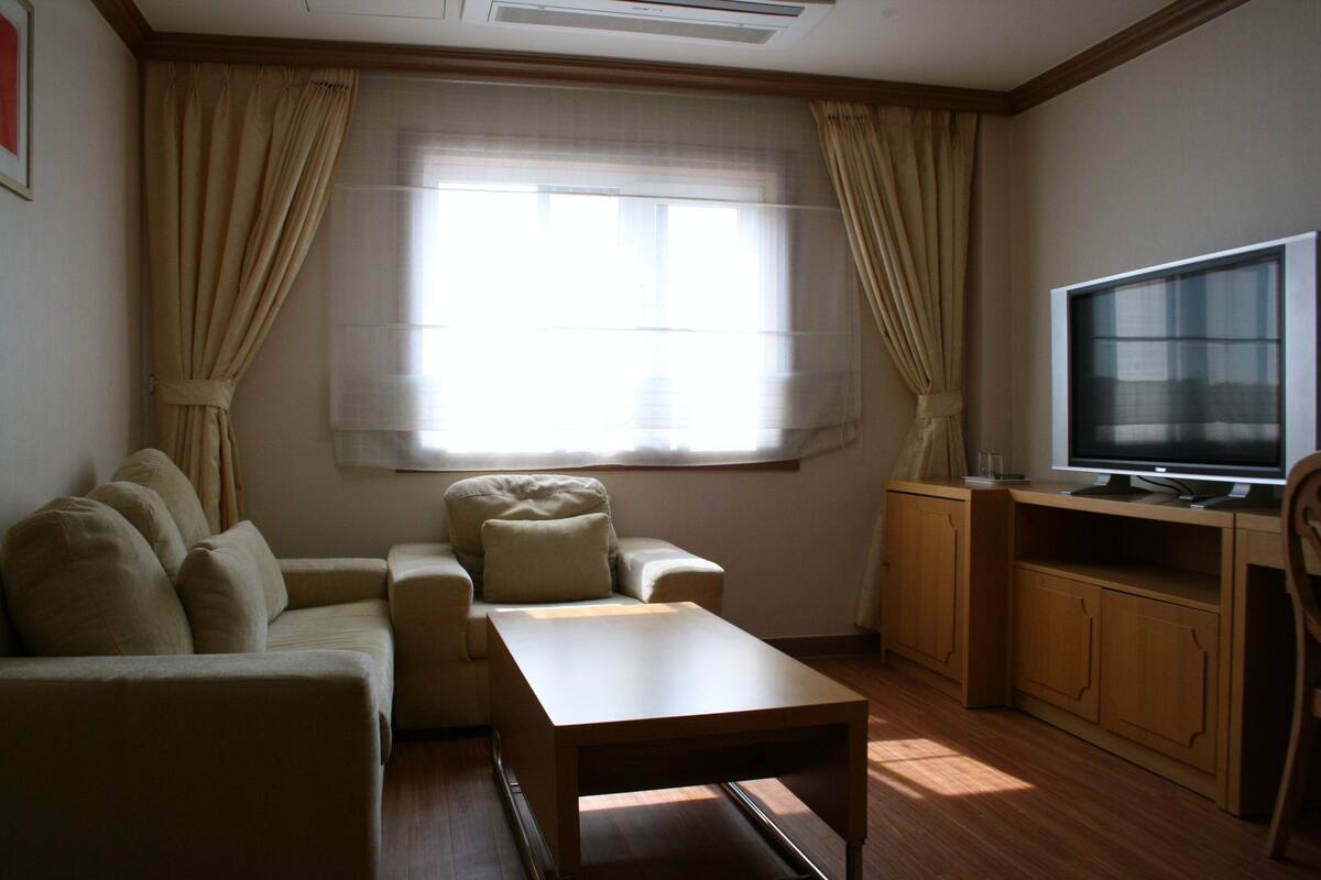 A living room with a sofa, a coffee table, a television on a wooden stand, and a window with curtains.
