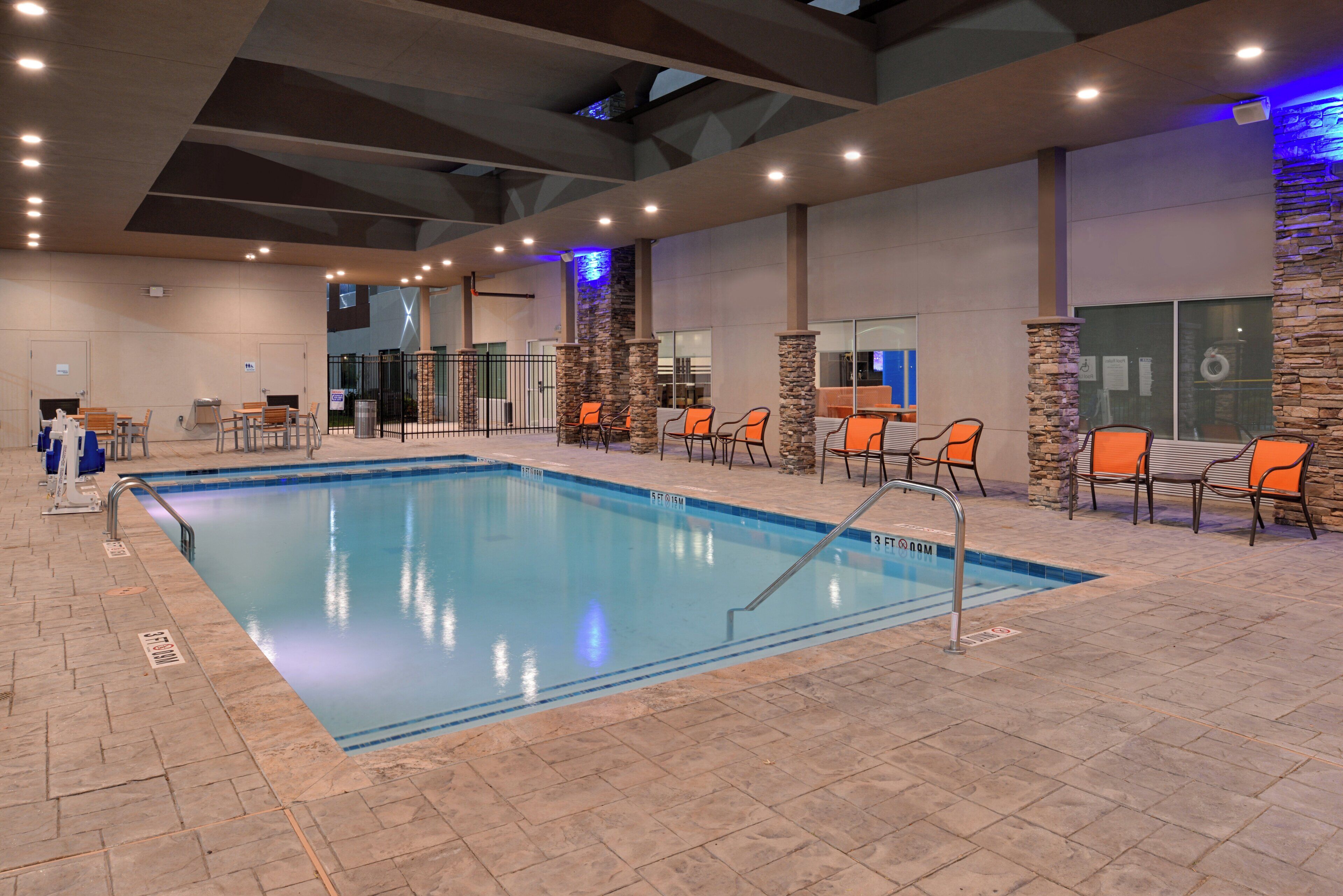 An indoor swimming pool with a tiled floor, surrounded by orange chairs and tables, and a stone wall on one side.