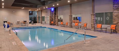 An indoor swimming pool with a tiled floor, surrounded by orange chairs and tables, and a stone wall on one side.