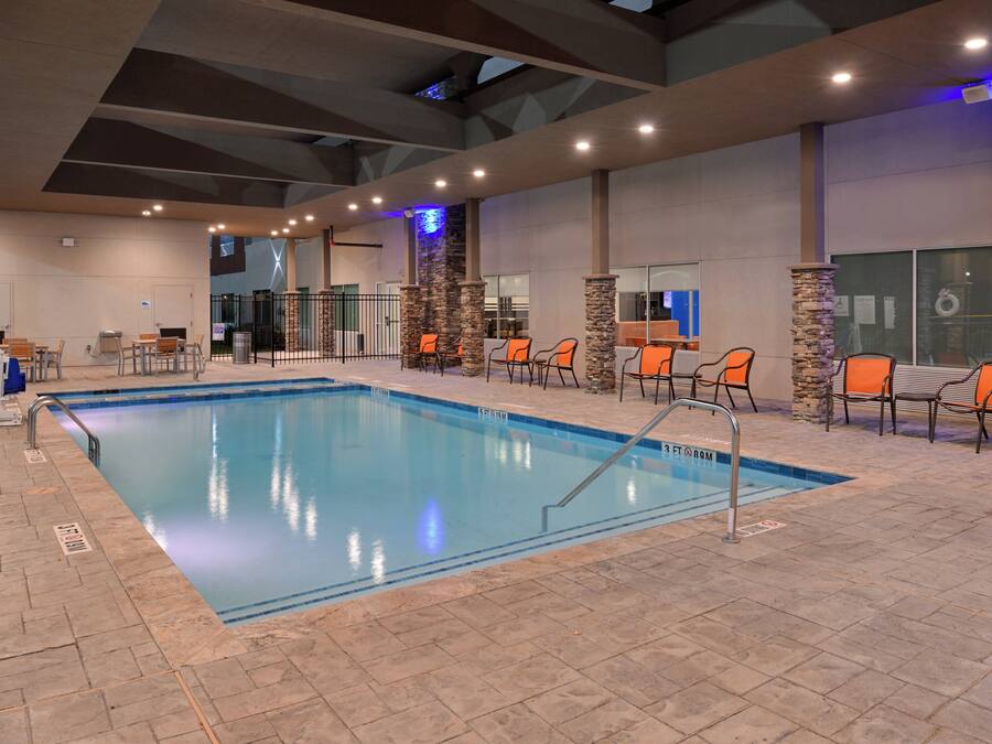 Una piscina interior con suelo de baldosas, rodeada de sillas y mesas naranjas, y una pared de piedra en un lateral.