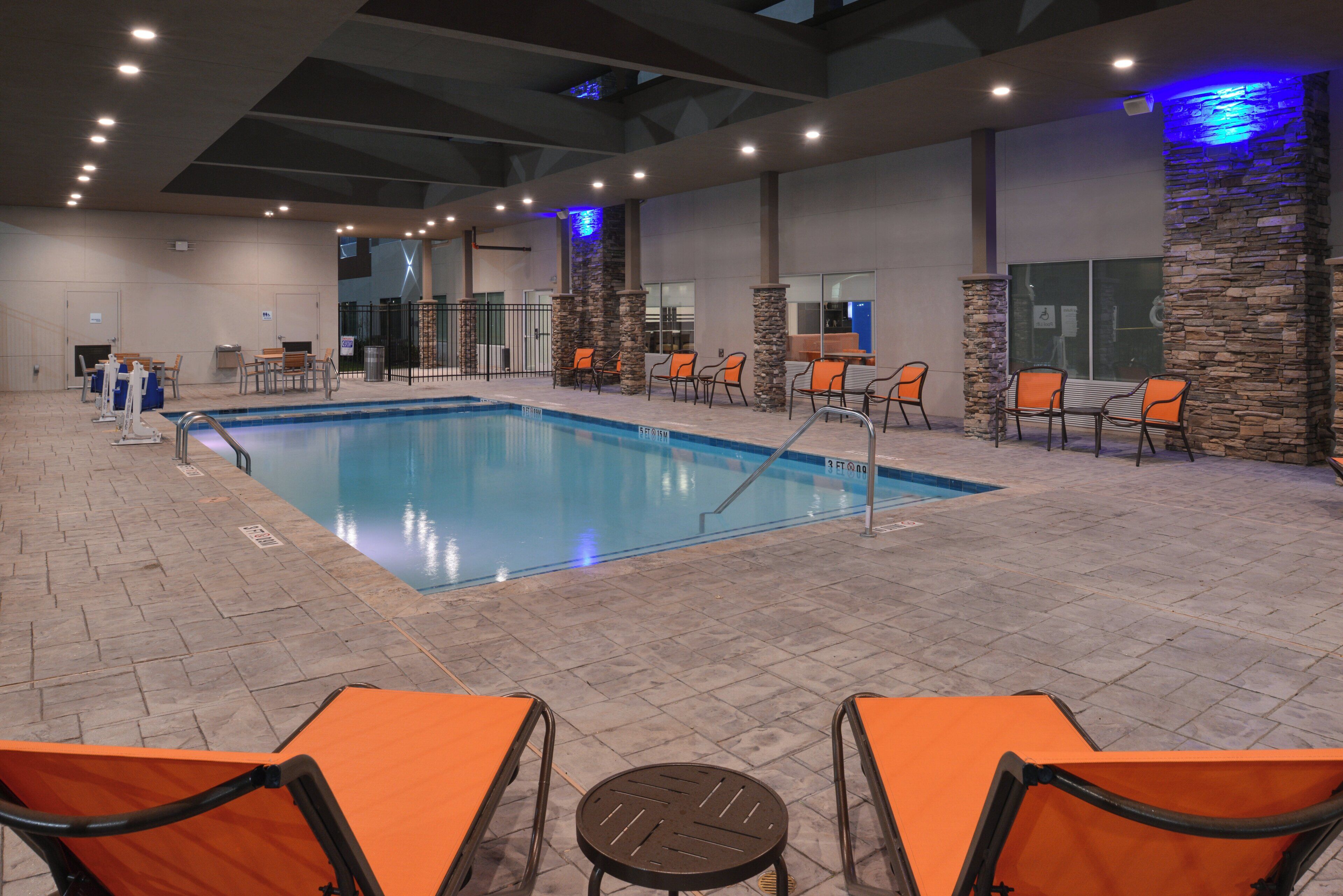 An indoor swimming pool with a tiled floor, surrounded by orange chairs and tables, and a stone wall with a blue light feature.