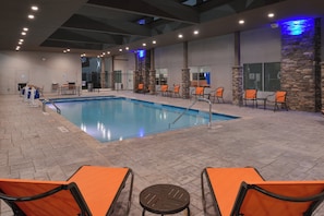 An indoor swimming pool with a tiled floor, surrounded by orange chairs and tables, and a stone wall with a blue light feature. - Holiday Inn Express & Suites Houston E - Pasadena by IHG (Pasadena)
