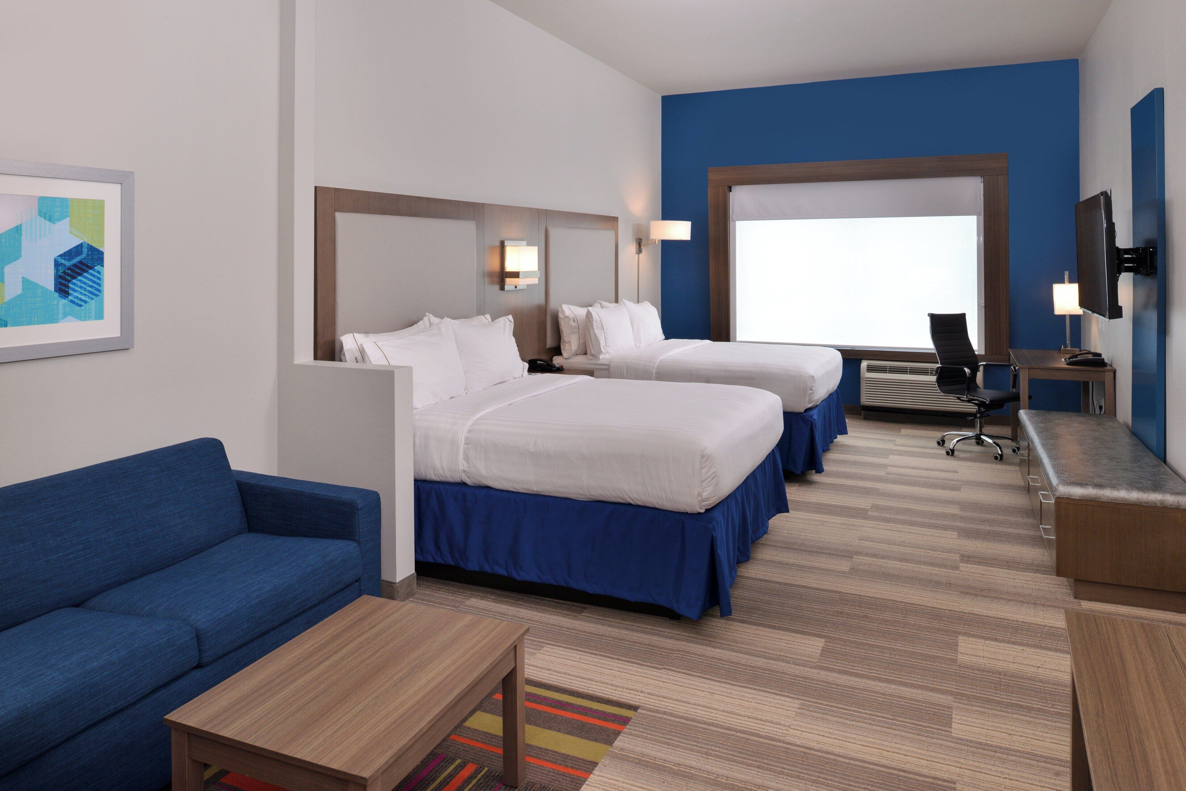 Habitación de hotel con dos camas, un sofá azul, una mesita, un escritorio con una silla y un televisor.