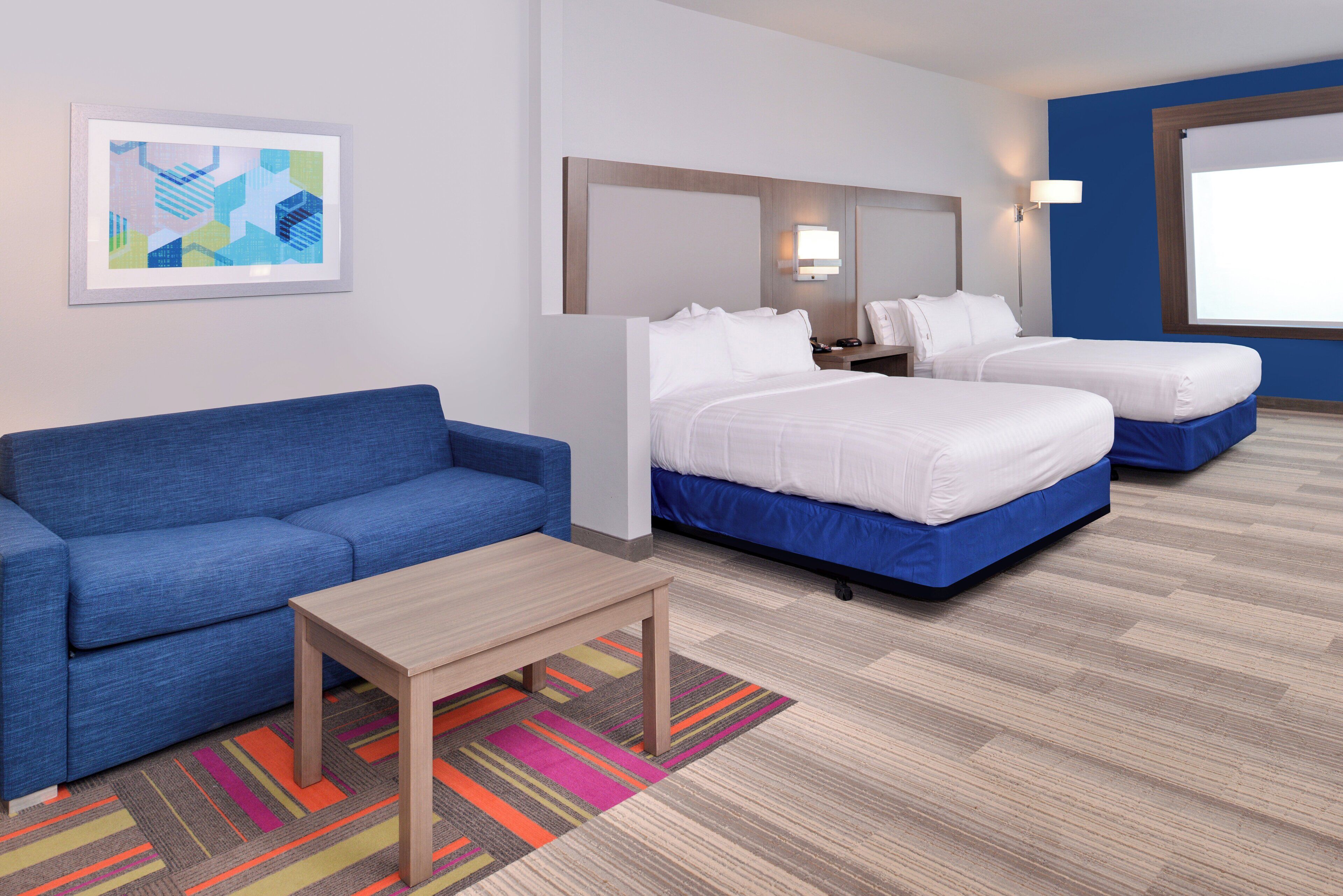 A hotel room with a blue sofa, two single beds, a small table, a colorful rug, and a framed abstract artwork on the wall.