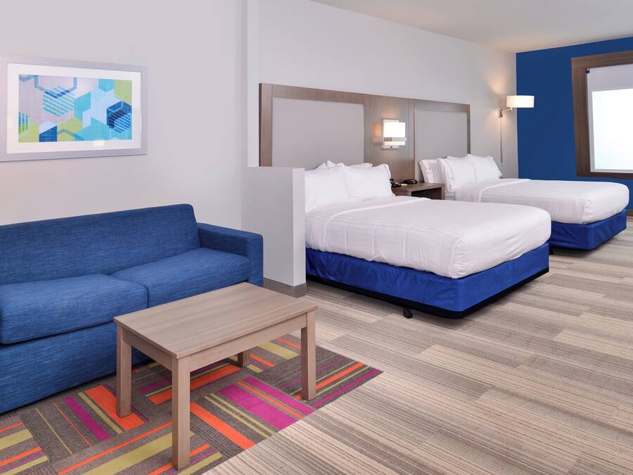 A hotel room with a blue sofa, two single beds, a small table, a colorful rug, and a framed abstract artwork on the wall.
