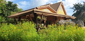 A traditional wooden house with a tiled roof, surrounded by lush greenery and yellow flowers.