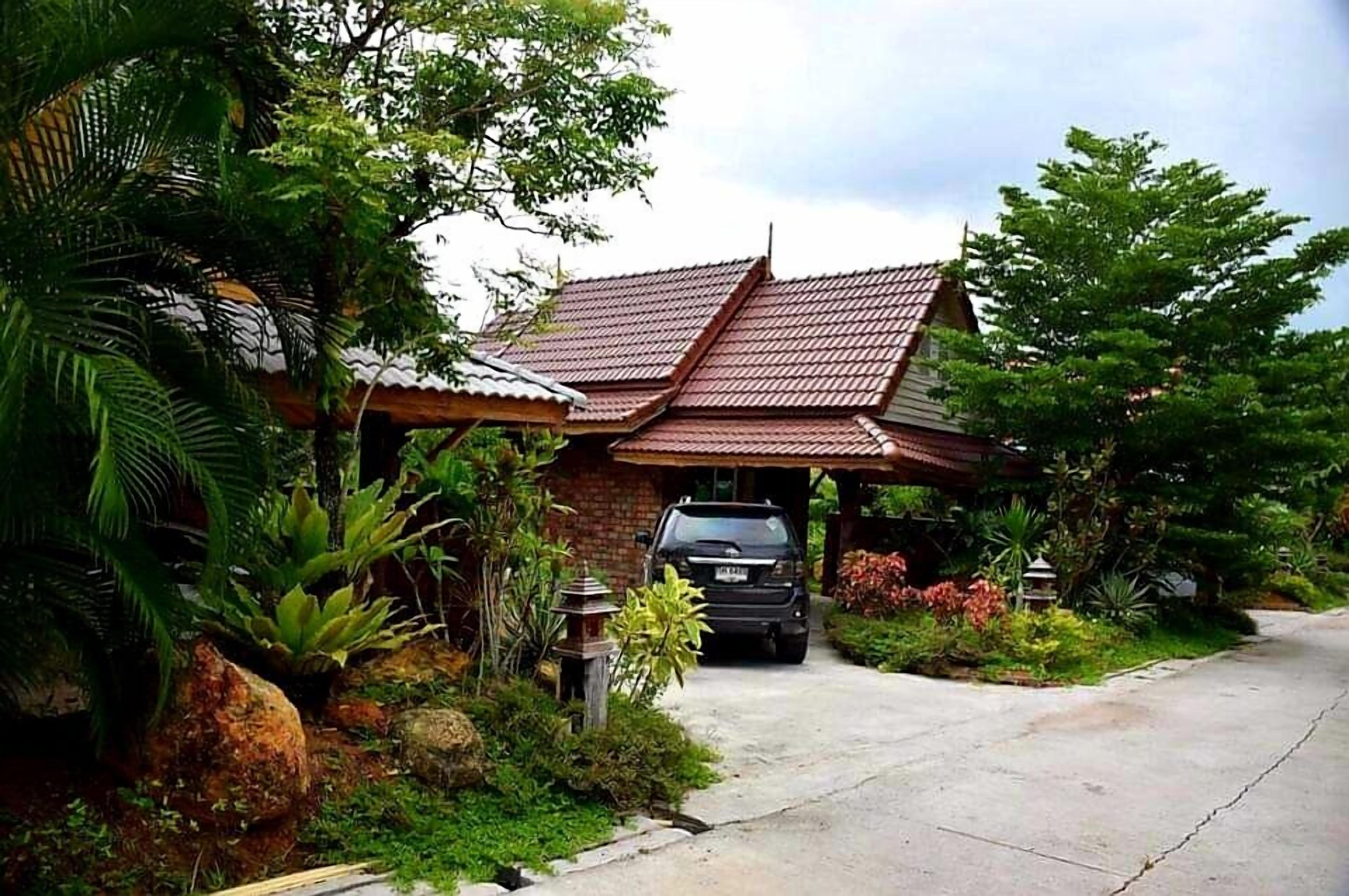 A traditional house with a tiled roof, surrounded by lush greenery and a paved driveway.