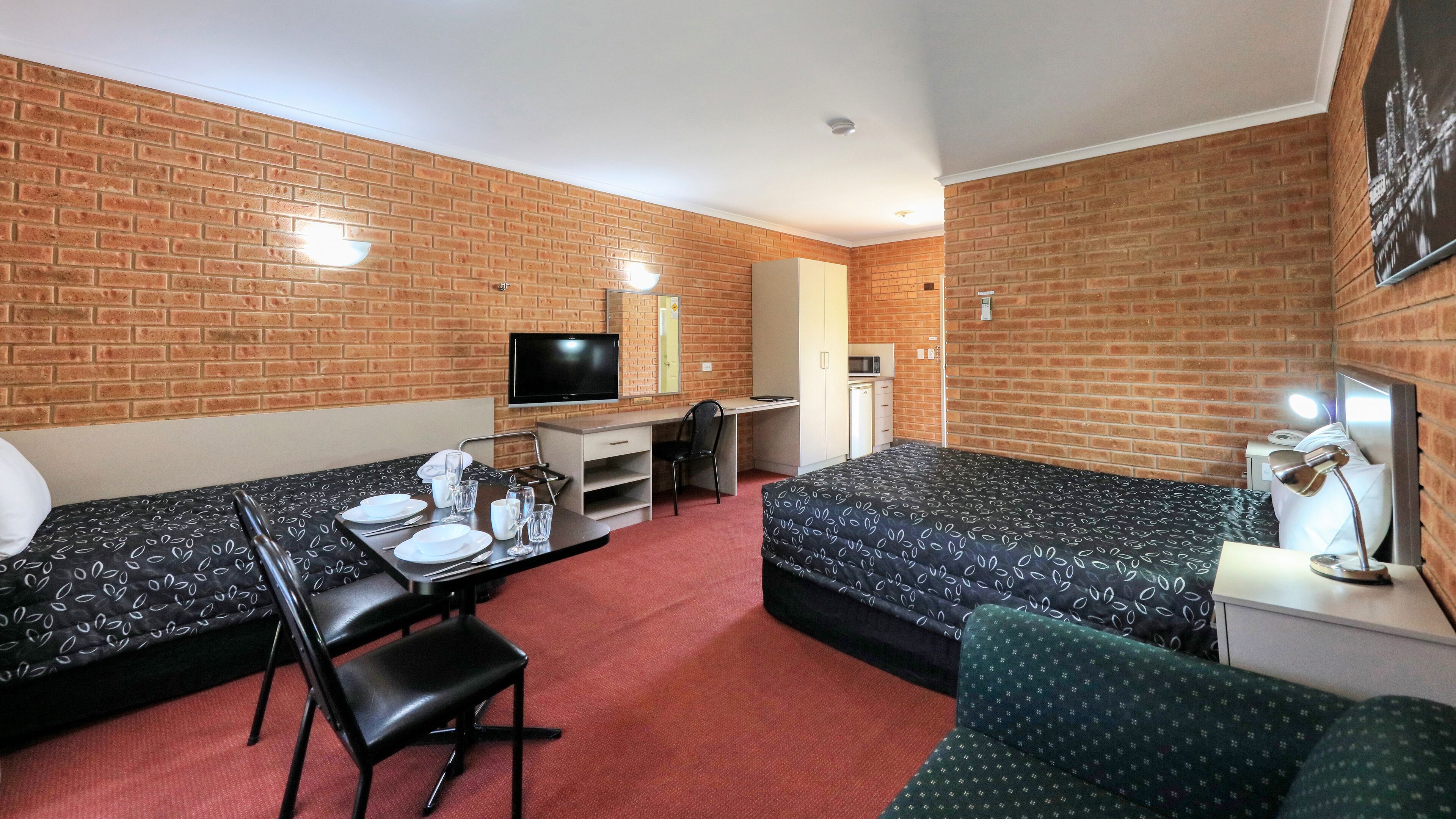 A hotel room with a brick wall, a bed, a desk with a chair, a TV, and a small kitchenette.