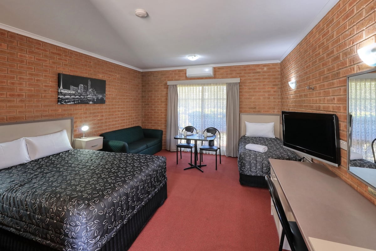 Une chambre d’hôtel avec un mur en briques, un lit recouvert d’une courtepointe à motifs, un bureau avec une chaise, un téléviseur et une fenêtre avec des rideaux.