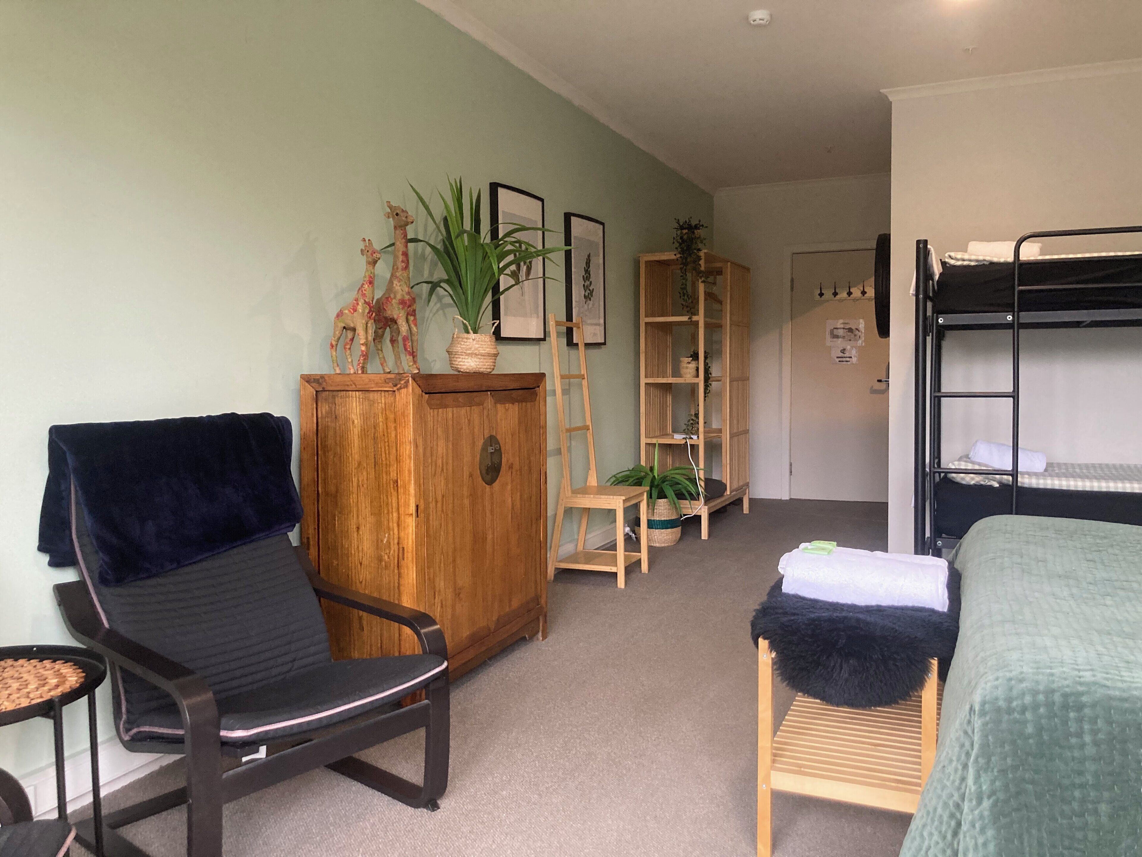 Habitación pequeña con literas, un armario de madera, una silla, una mesita y una planta.
