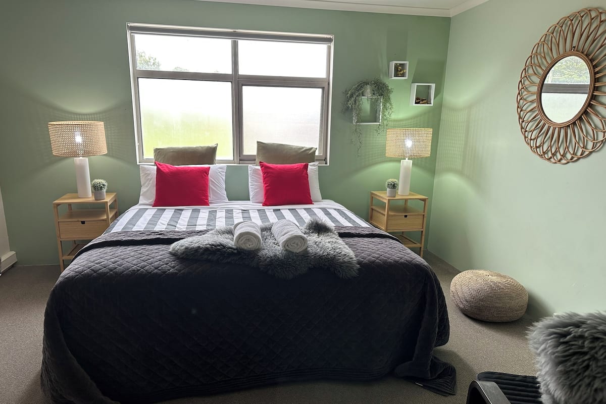 A neatly made bed with a striped duvet, two red pillows, and rolled towels. There are two bedside tables with lamps and a decorative mirror on the wall.