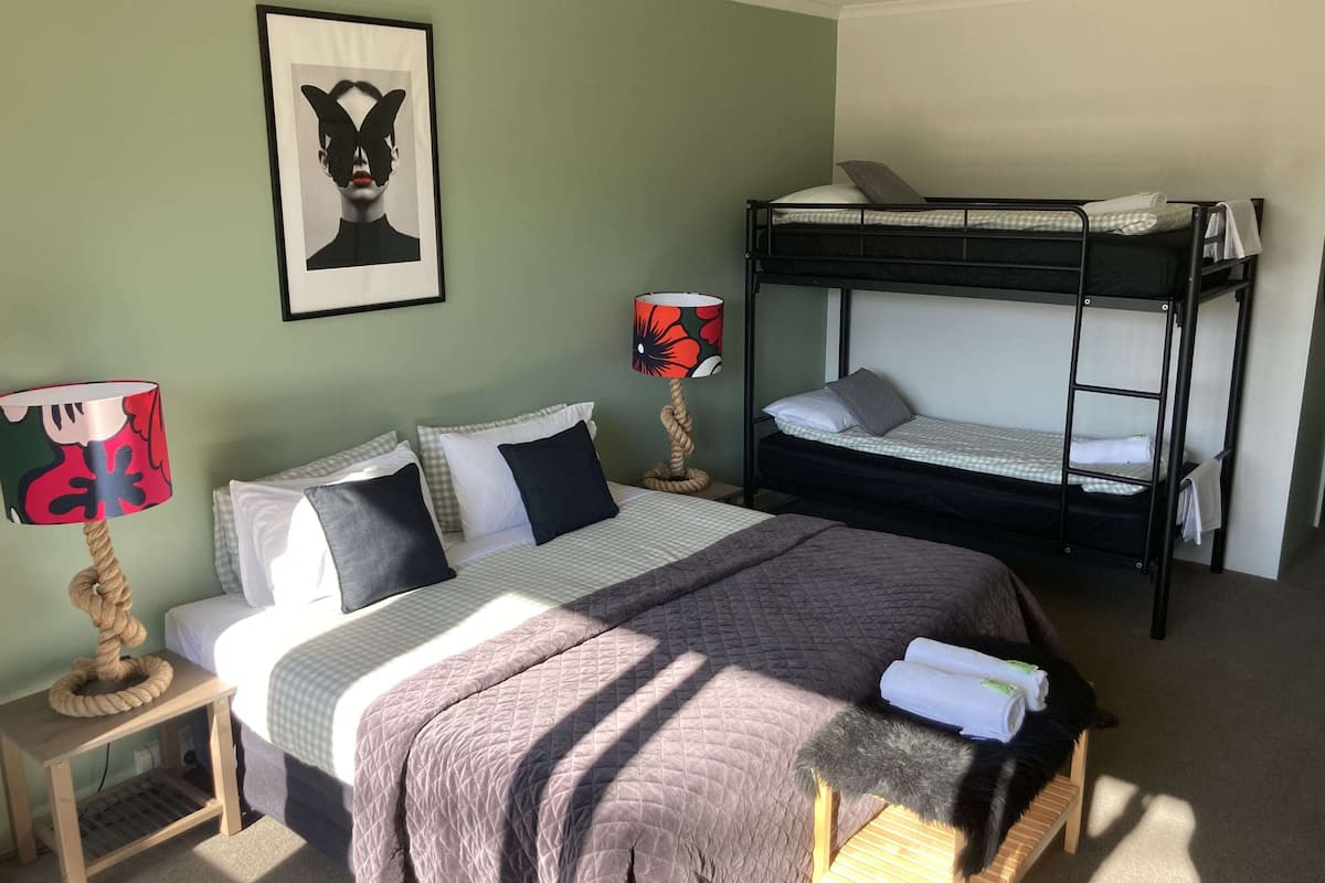 A room with two beds, one with a bunk and one with a standard bed. There are two lamps, a framed picture on the wall, and a cat on a chair.