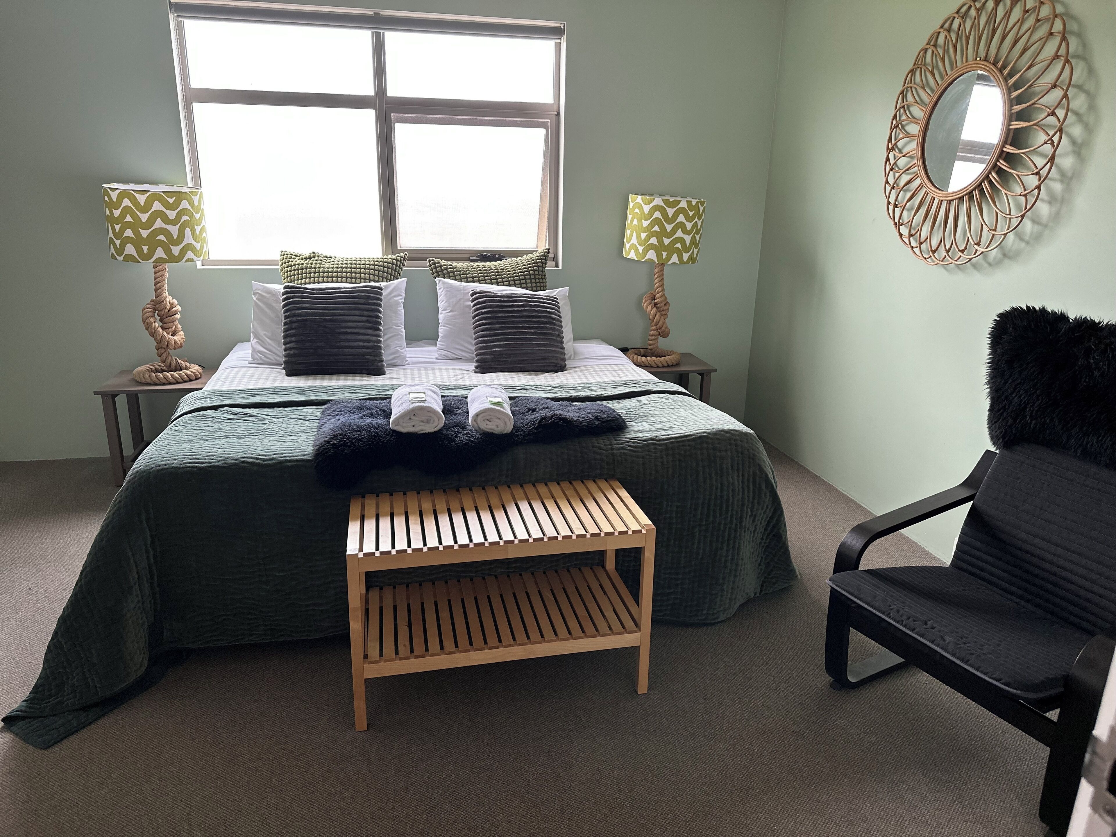 Een keurig opgeruimde slaapkamer met een bed, twee bedlampen, een houten bankje, een zwarte stoel en een decoratieve spiegel aan de muur.