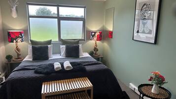 Un dormitorio ordenado con una cama grande, dos lámparas de noche, una ventana con vistas, un pequeño banco y un jarrón con flores.