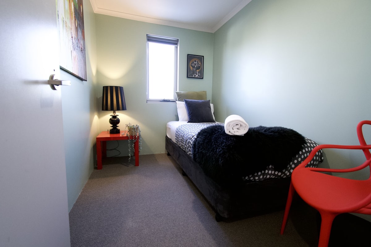 A bedroom with a bed, a window, a red chair, a small table with a lamp, and a picture on the wall.