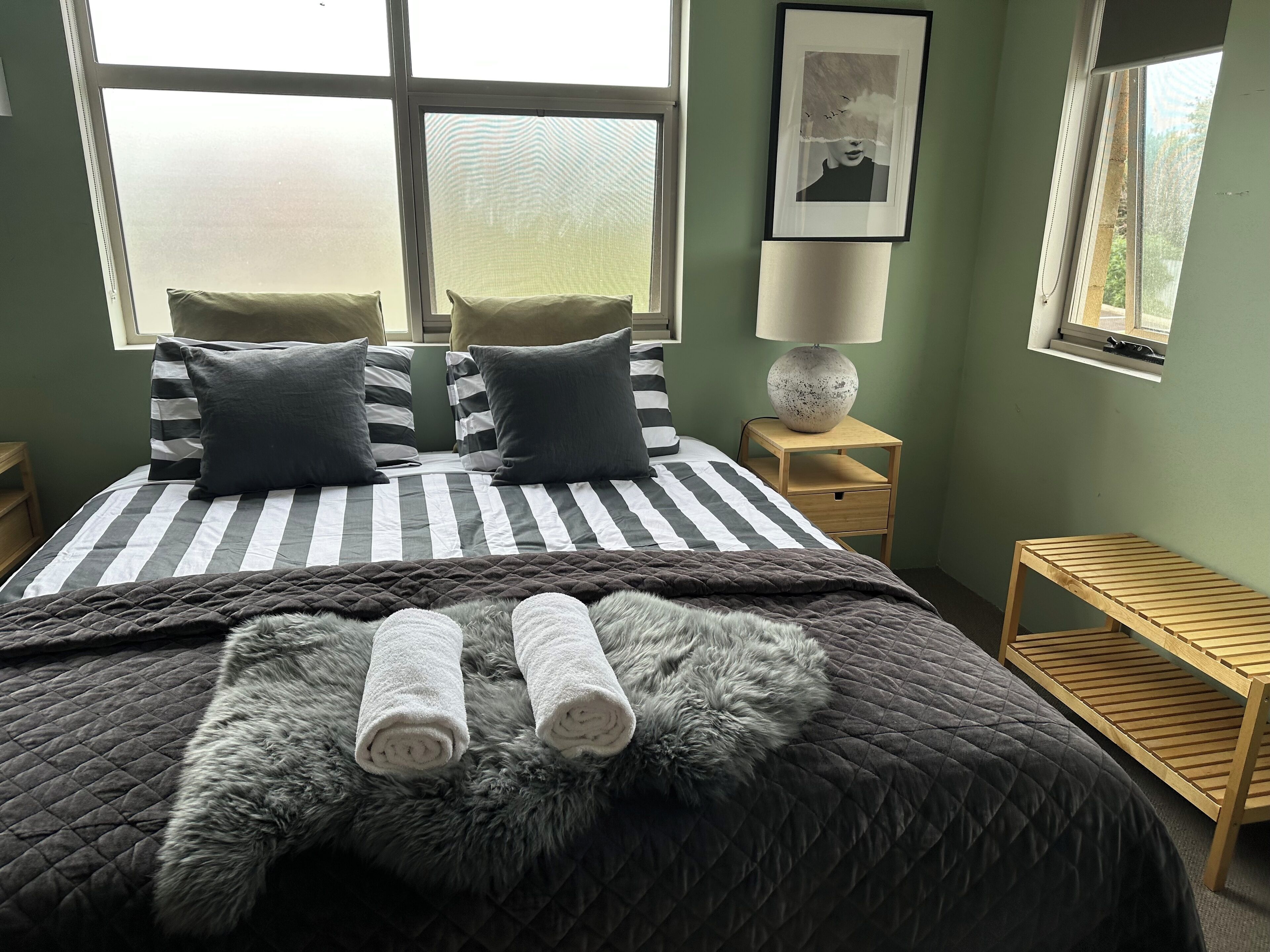 A neatly made bed with striped bedding and two pillows, a fluffy rug, and two rolled towels.