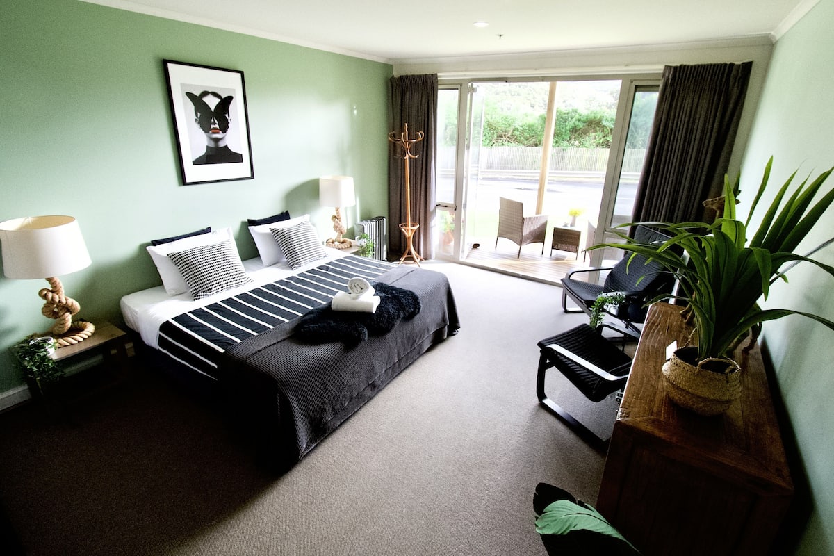 A hotel room with a bed, a chair, a table, a lamp, a plant, and a view of the outdoors.
