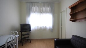 Uma sala de estar com televisão em um móvel, um sofá preto, uma mesa de jantar com cadeiras e uma janela com cortinas leves.