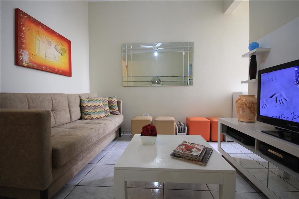 A living room with a beige sofa, a white coffee table, a flat-screen TV, a glass shelf with decorative items, and a painting on the wall.