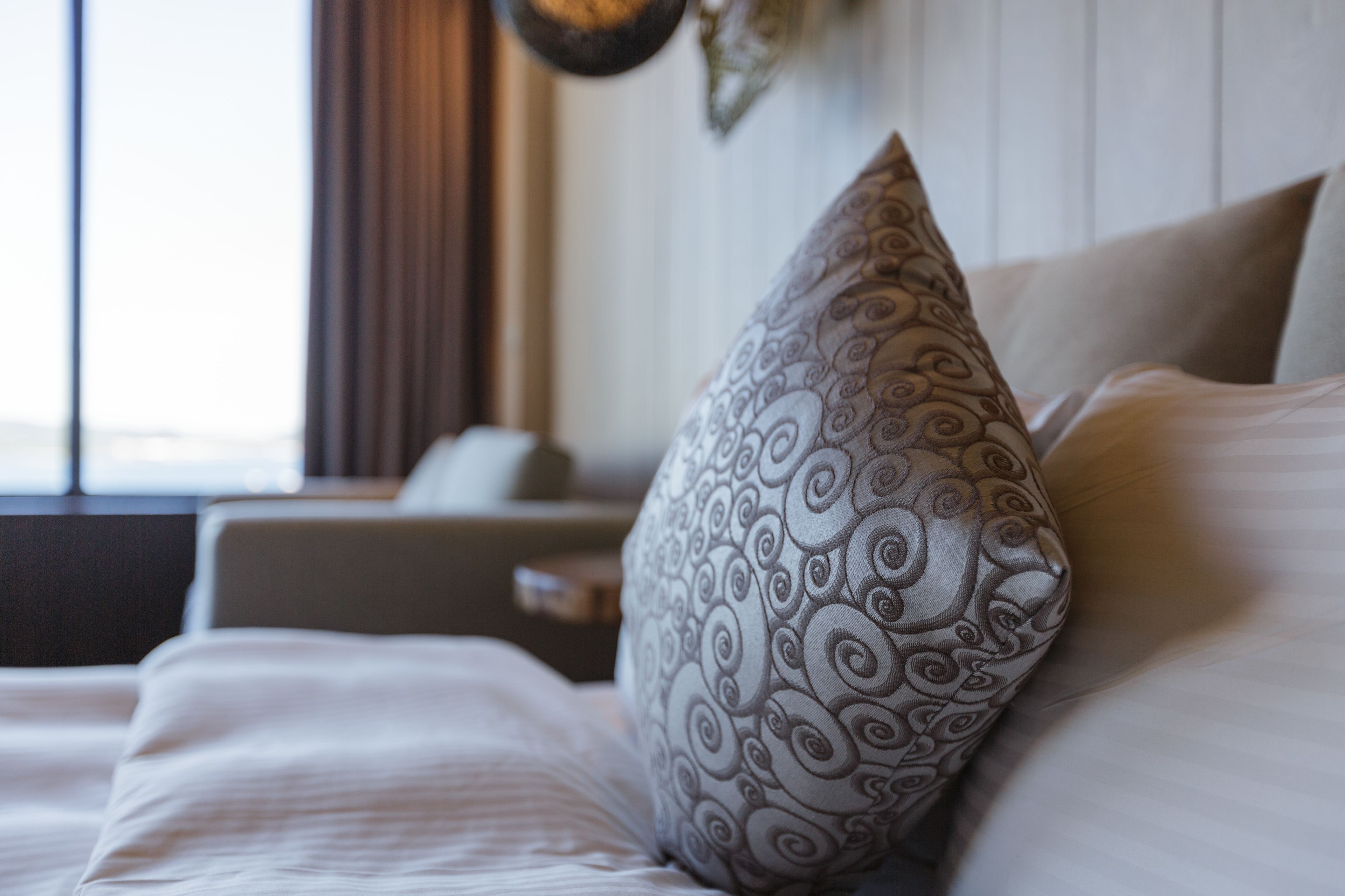 A neatly made bed with a patterned pillow and a plain pillow, a bedside table, and a window with curtains.