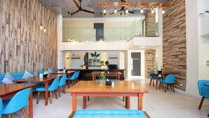 Een ruime eethoek met houten tafels en blauwe stoelen, een centrale bank, en een keuken op de achtergrond.