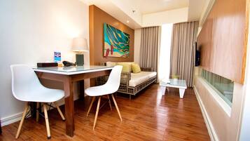 Habitació d'hotel moderna amb sòl de fusta, zona de menjador amb taula de vidre i cadires blanques, sofà amb coixins, televisió penjada a la paret i un quadre abstracte gran sobre el sofà.