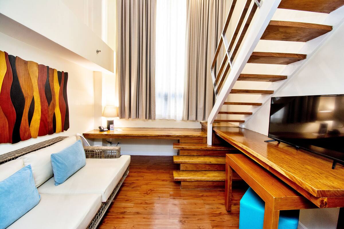 A modern living room with a wooden staircase, a sofa with blue cushions, and a flat-screen TV on a wooden table.