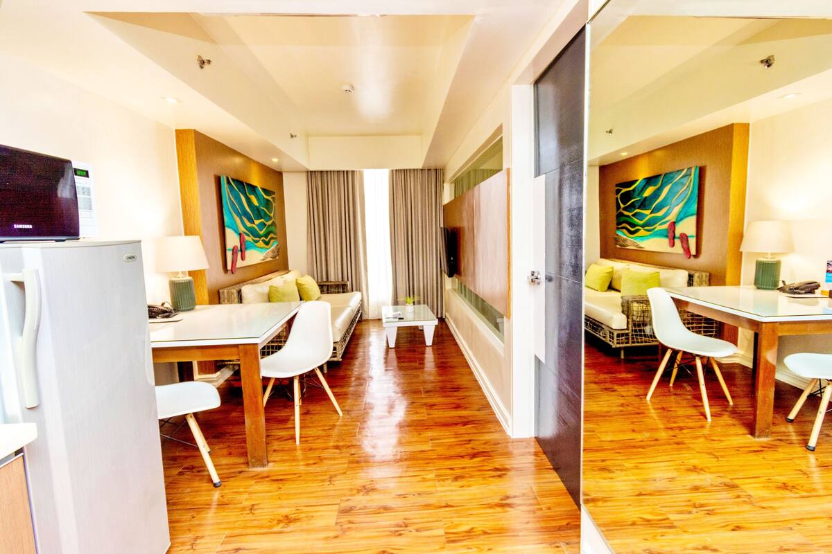 A hotel room with a wooden floor, a sofa with cushions, a dining table with chairs, a TV, and a painting on the wall.
