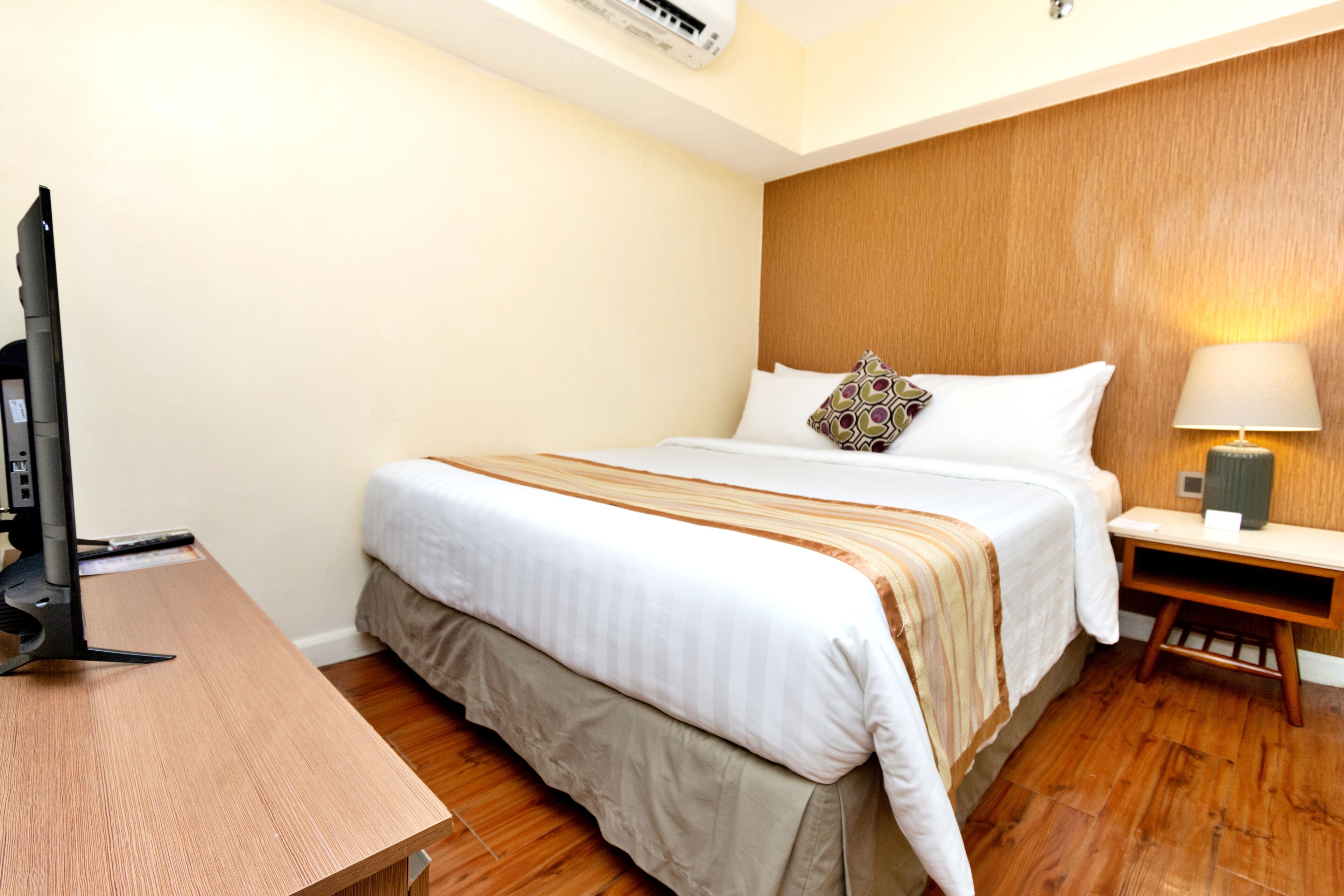 A hotel room with a bed, a desk, a TV, a lamp, and a wooden headboard.