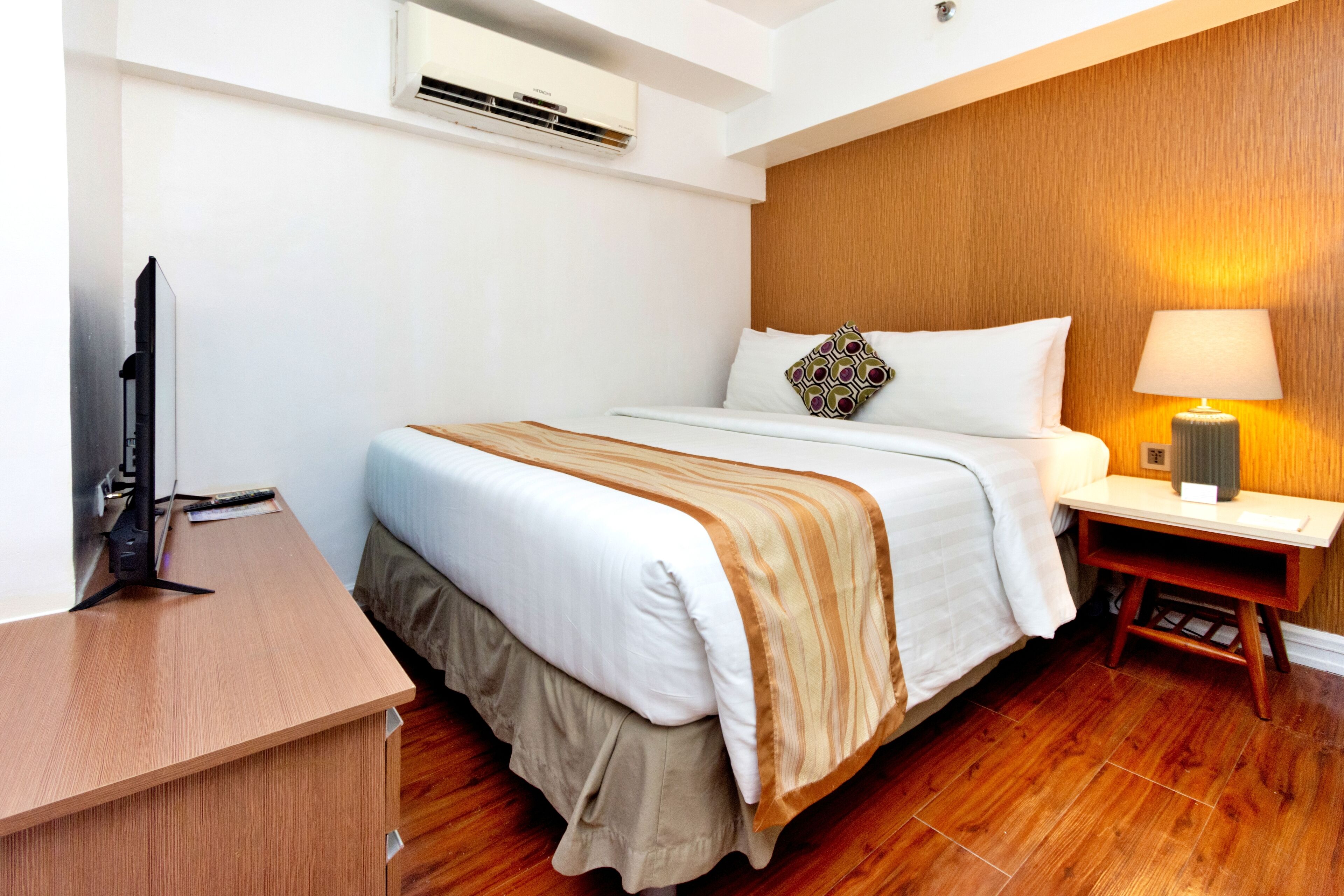 A hotel room with a wooden floor, a bed with a white and beige bedspread, a bedside table with a lamp, and a flat-screen TV on a wooden desk.