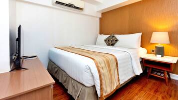A hotel room with a wooden floor, a bed with a white and beige bedspread, a bedside table with a lamp, and a flat-screen TV on a wooden desk.