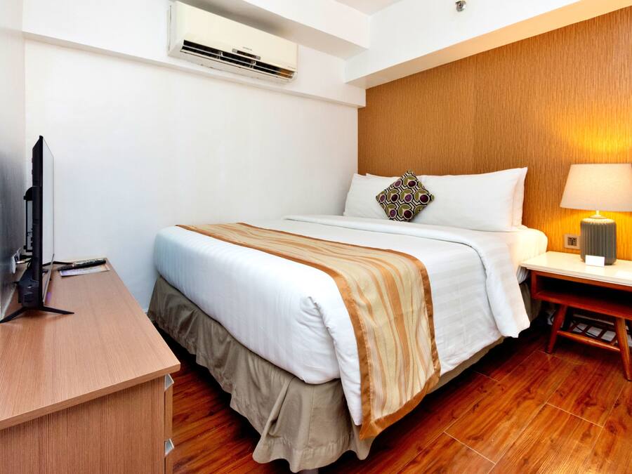 A hotel room with a wooden floor, a bed with a white and beige bedspread, a bedside table with a lamp, and a flat-screen TV on a wooden desk.