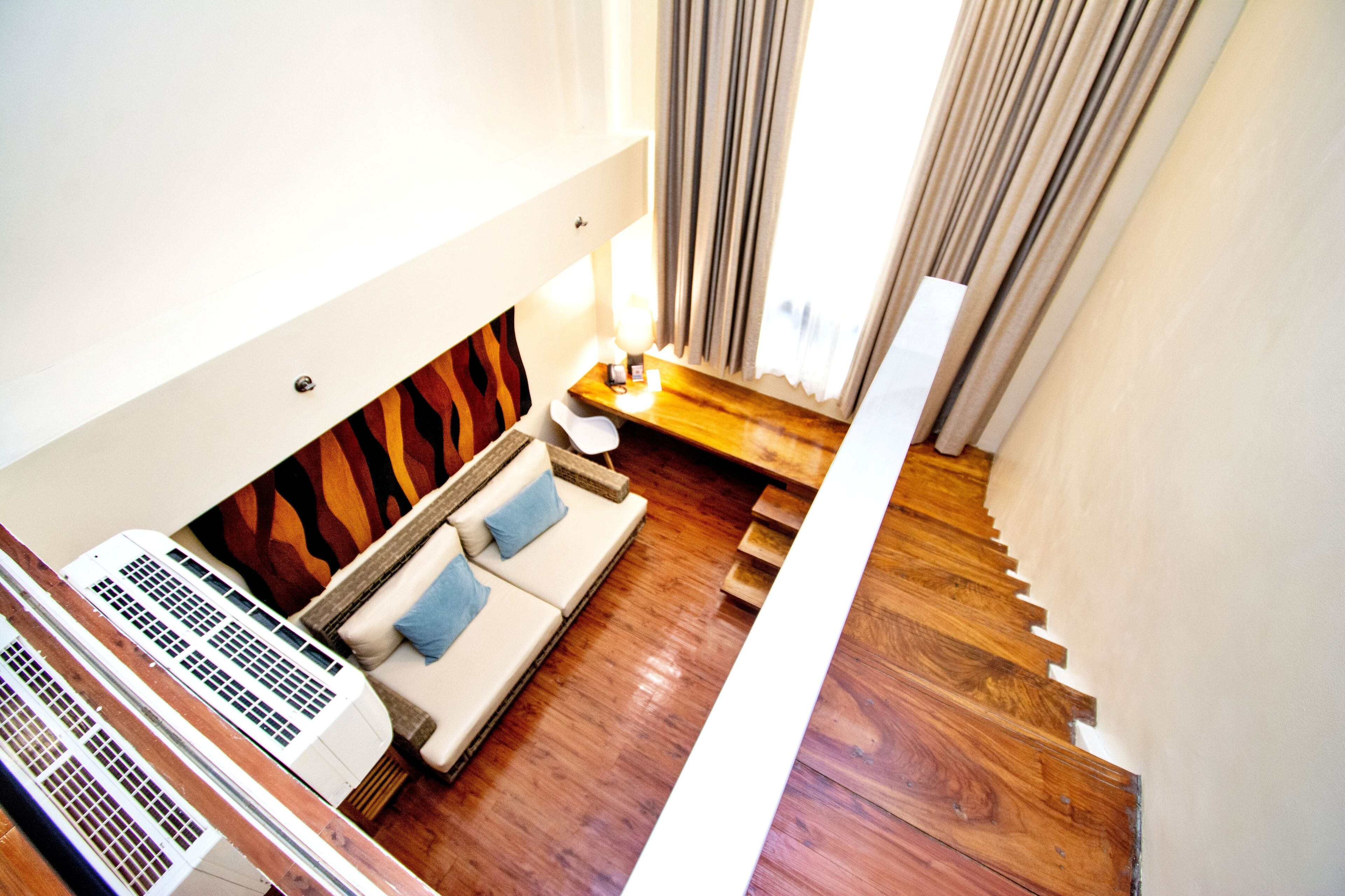 Una habitación de hotel moderna con suelo de madera, un sofá con cojines azules, una mesita con lámpara y una escalera.