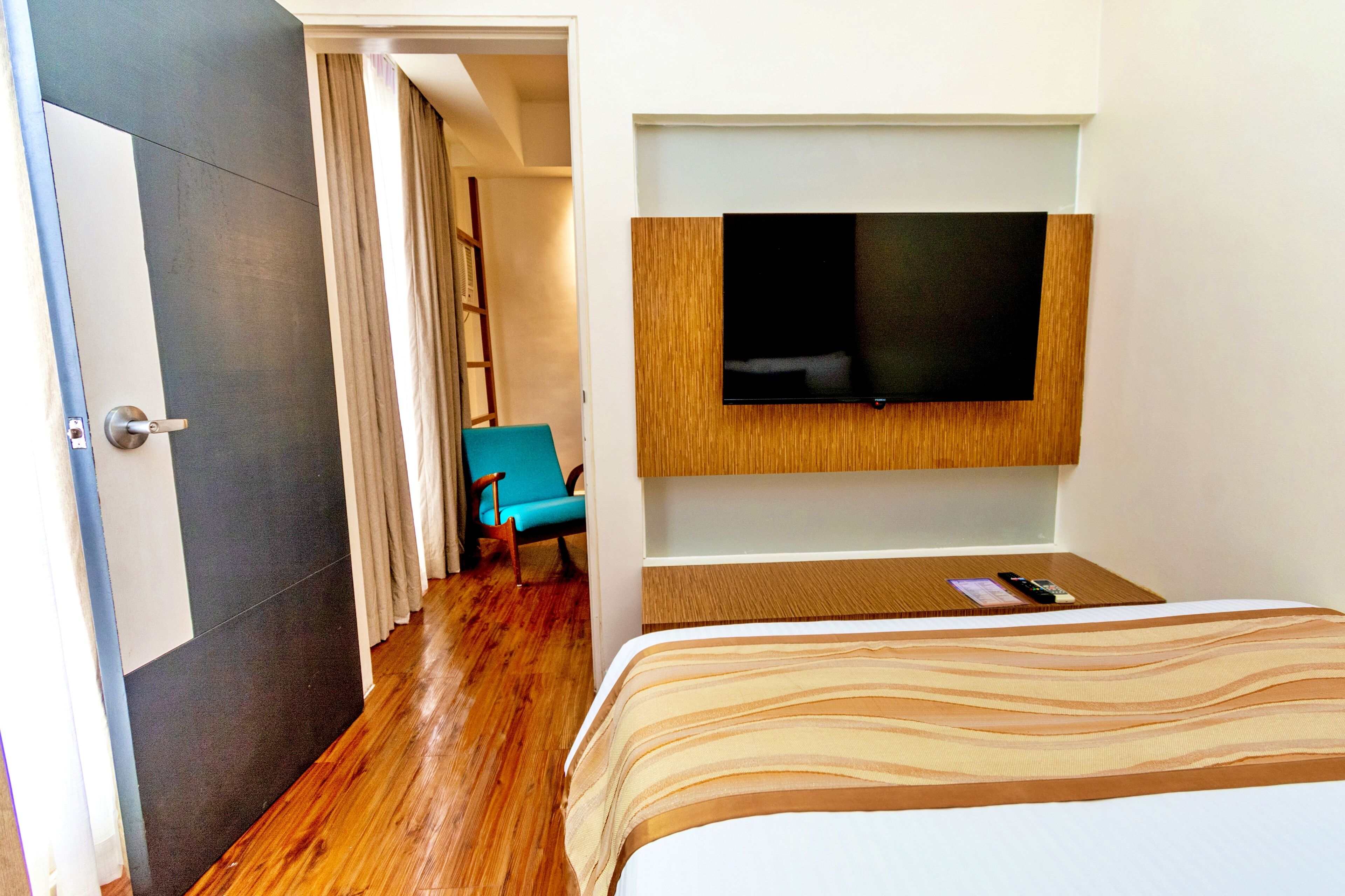 Une chambre d’hôtel avec un lit, un téléviseur fixé au mur, un plancher en bois, une chaise bleue et une porte.