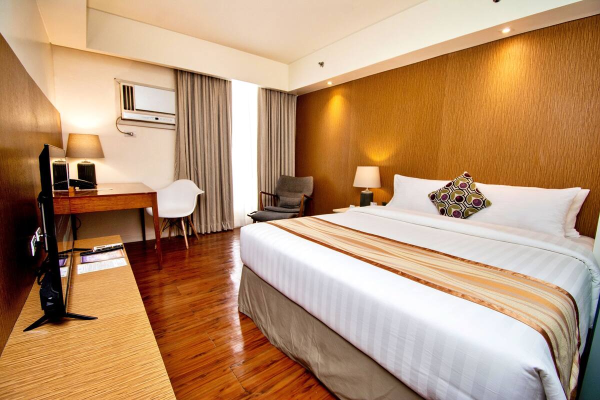 A hotel room with a wooden floor, a bed with white bedding and a patterned pillow, a desk with a computer and lamp, and a chair.