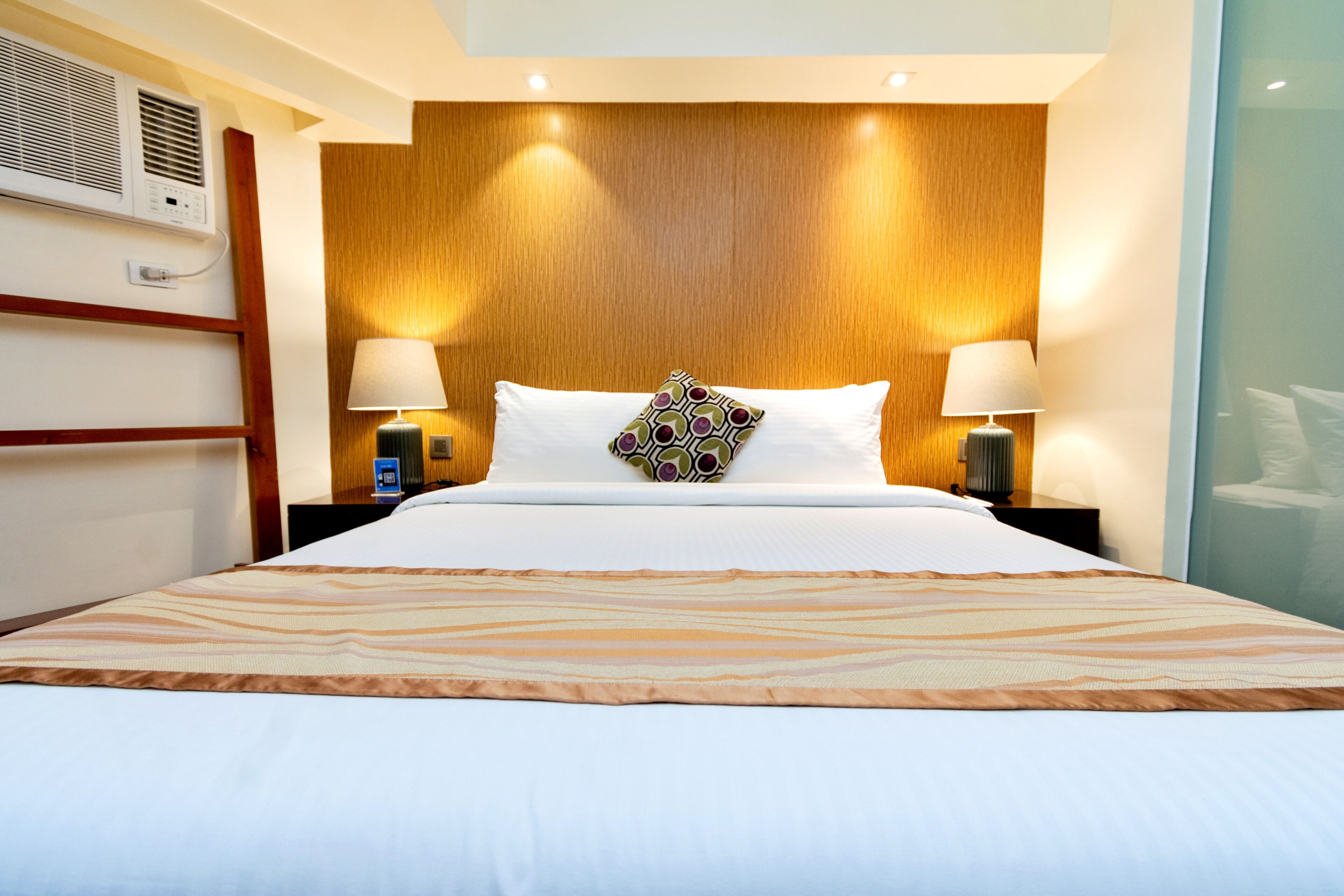 Una habitación de hotel con una cama grande, dos lámparas de noche, un cabecero de madera y un aire acondicionado instalado en la pared.