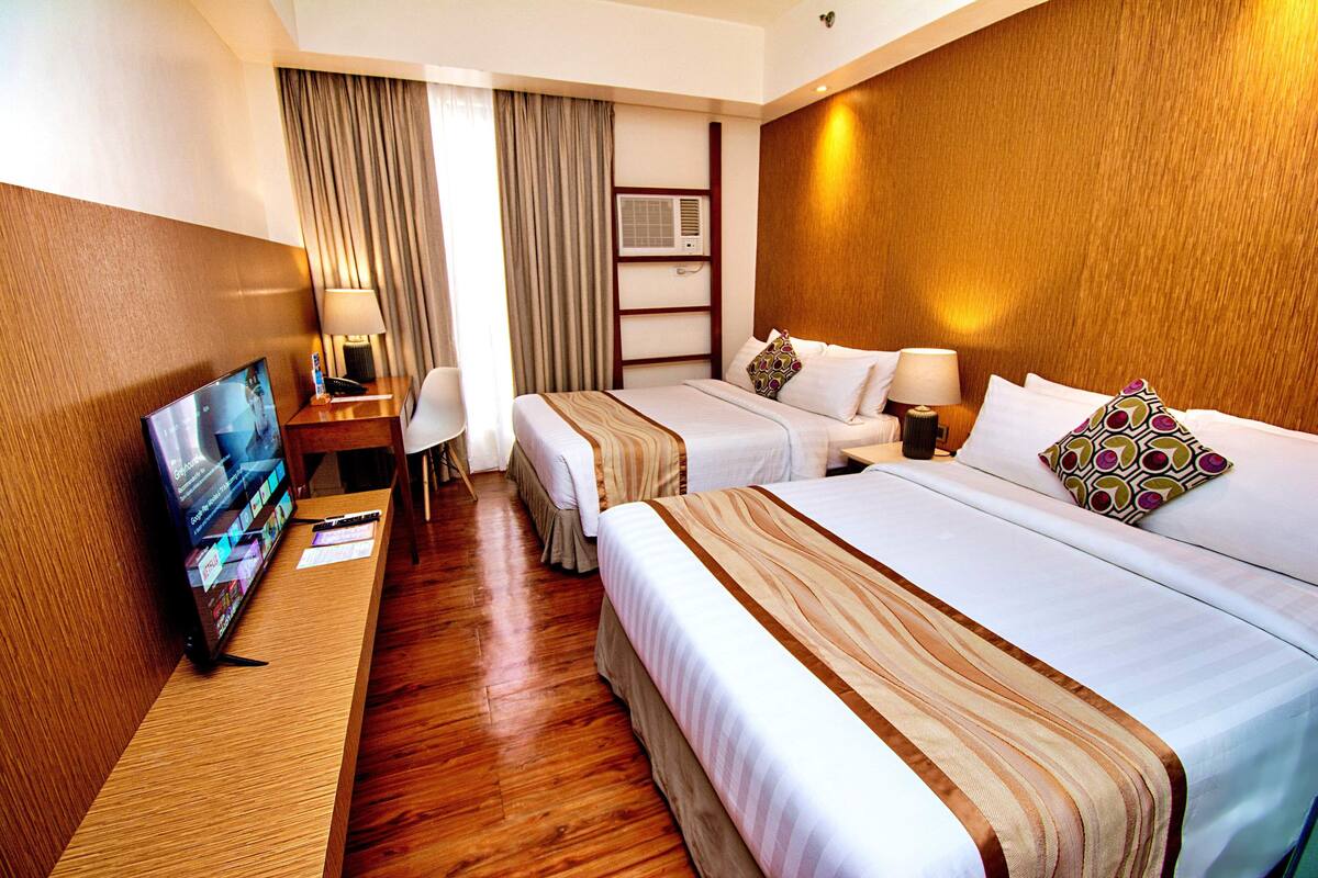 A hotel room with a wooden floor, a TV on a wooden stand, two beds with white linens and striped blankets, a desk with a lamp, and a window with curtains.