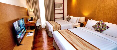 Habitación de hotel con piso de madera, un televisor sobre un soporte de madera, dos camas con sábanas blancas y colchas a rayas, un escritorio con lámpara y una ventana con cortinas.