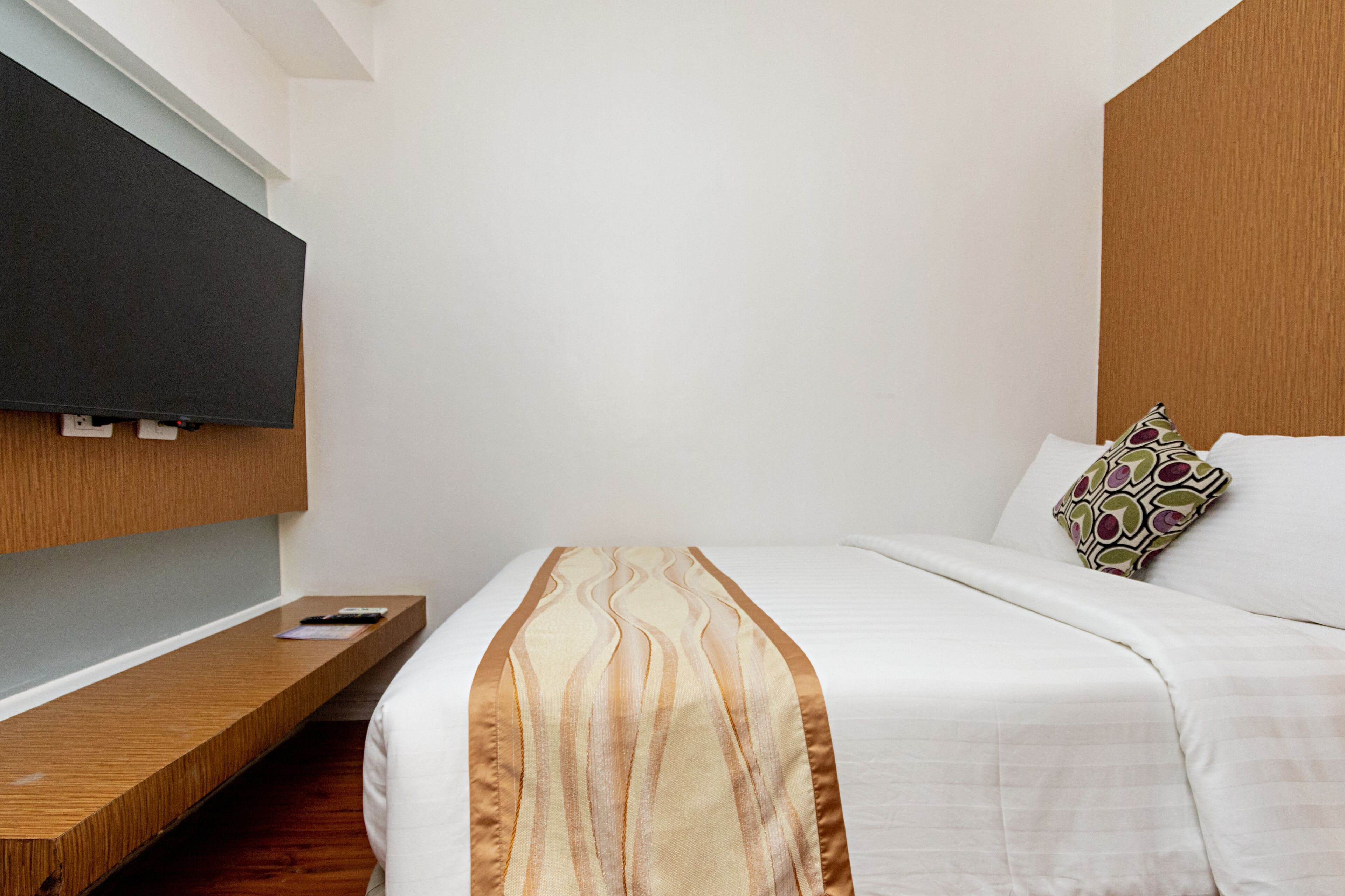 Une chambre d’hôtel avec un lit, un téléviseur fixé au mur, une table de chevet en bois et un coussin à motifs.