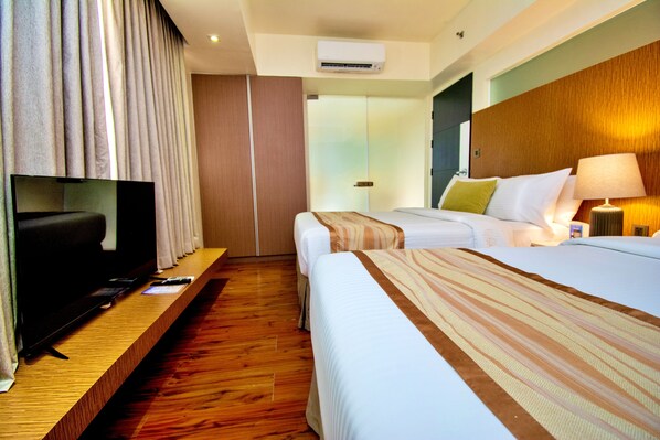 A hotel room with a flat-screen TV on a wooden shelf, two beds with striped bedding, a wooden headboard, and a bedside lamp.