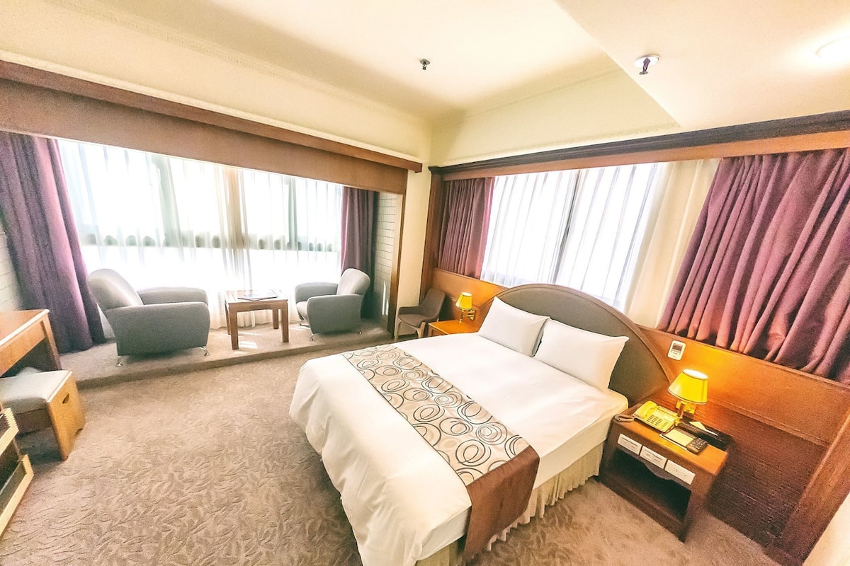 Habitación de hotel con una cama grande, dos sillones, una mesita y una mesita de noche con lámpara.