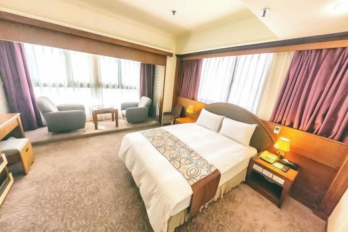 A hotel room with a large bed, a desk, a chair, a small table, and a view of the outdoors.