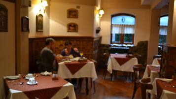 Um restaurante com mesas cobertas por toalhas brancas e jogos americanos vermelhos, cadeiras de madeira e luminárias fixadas na parede.