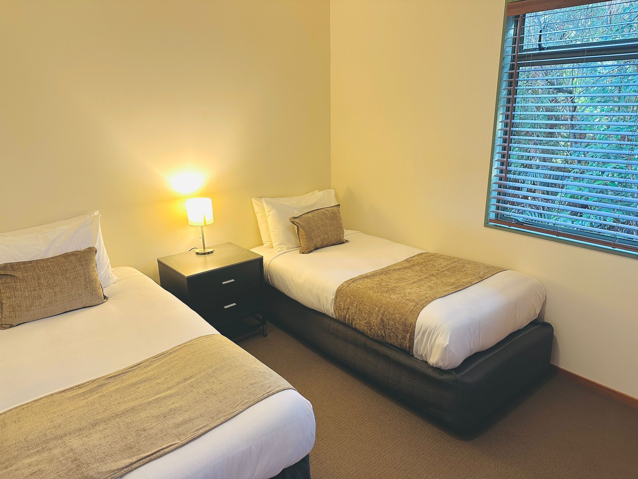 Habitación de hotel con dos camas individuales, una mesita de noche con lámpara y una ventana con persianas.