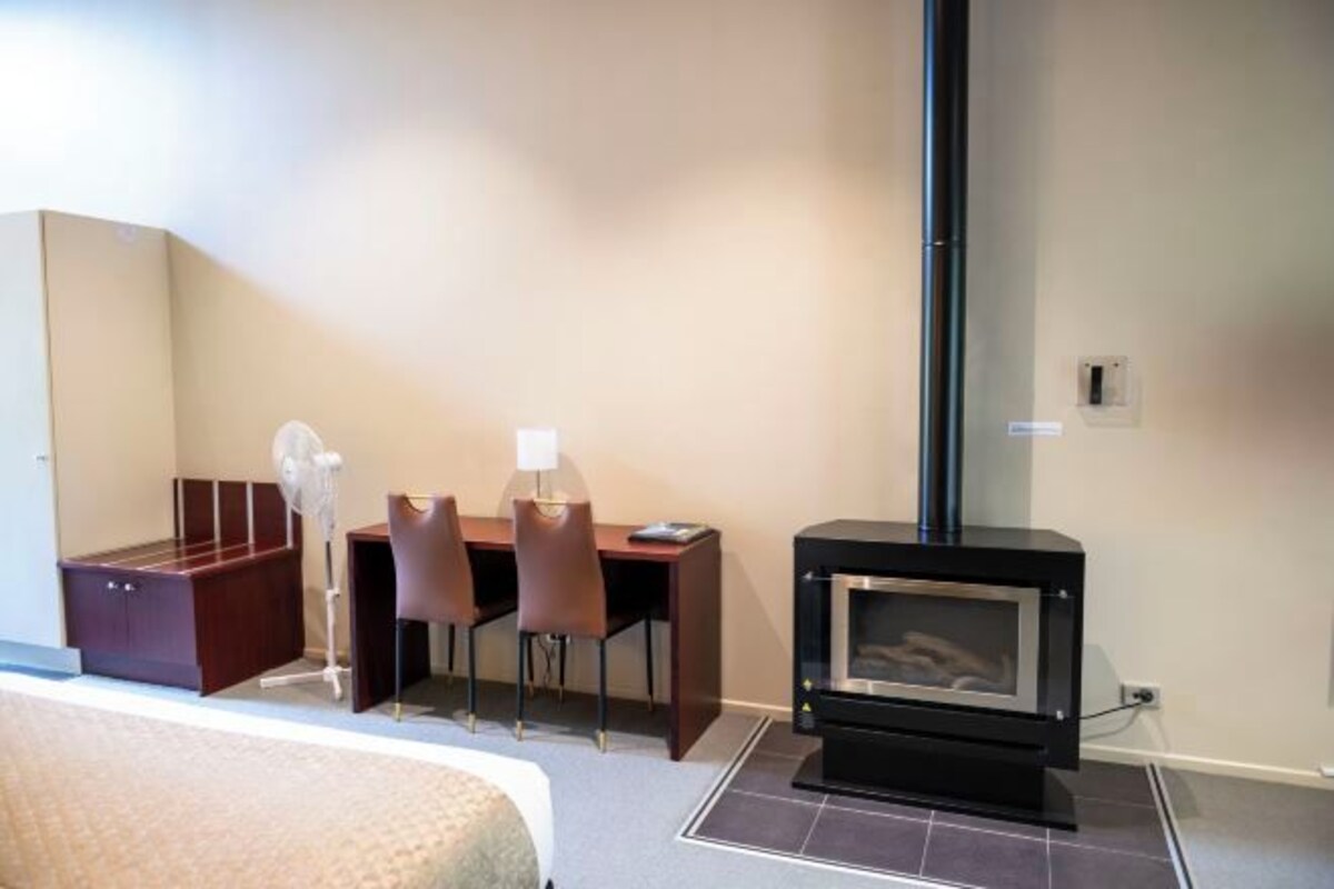A hotel room with a bed, a desk with chairs, a small table, a lamp, and a wood-burning stove.