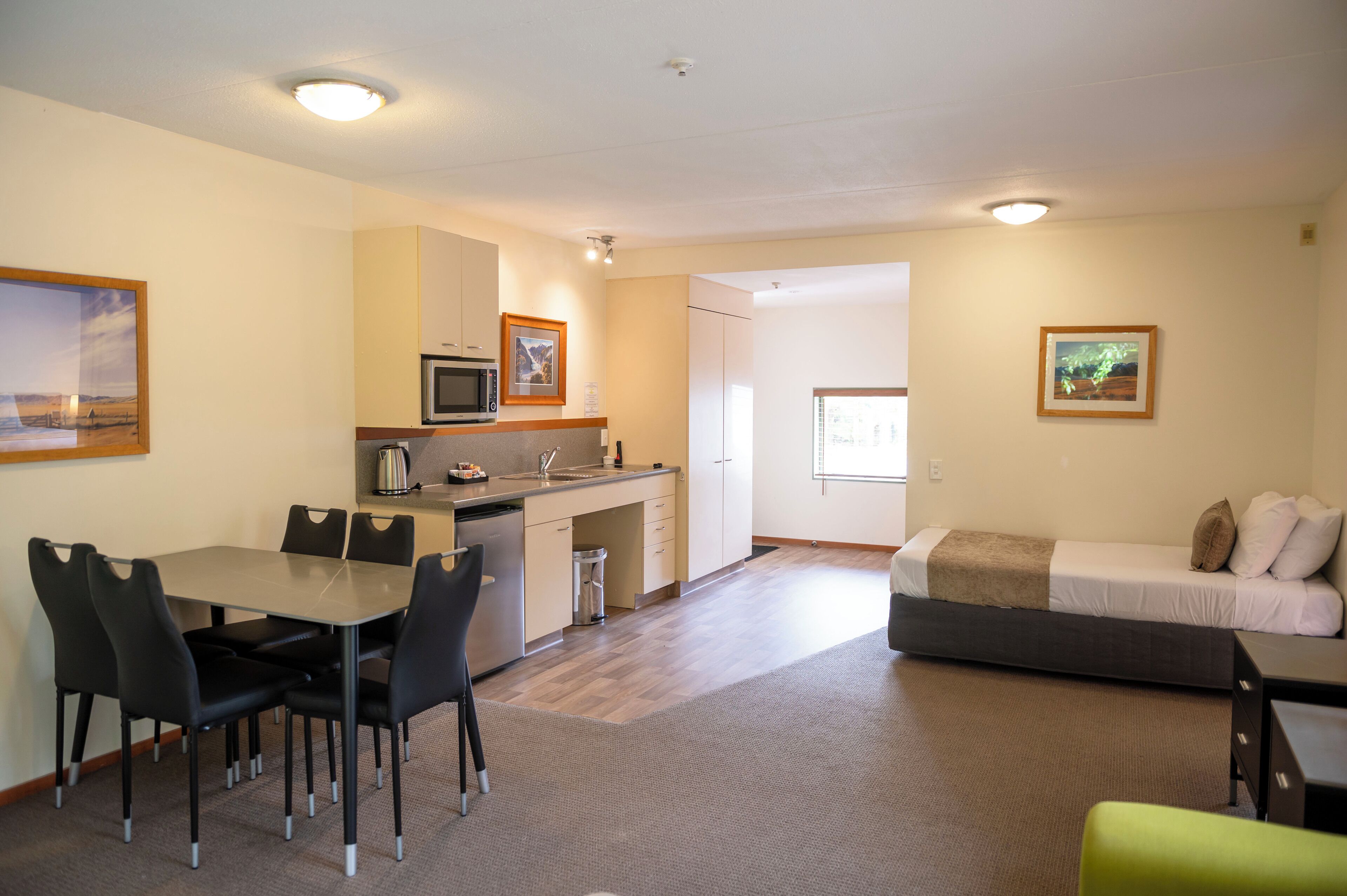 A compact hotel room with a kitchenette, a dining area with a table and chairs, a bed with pillows, and a small desk.