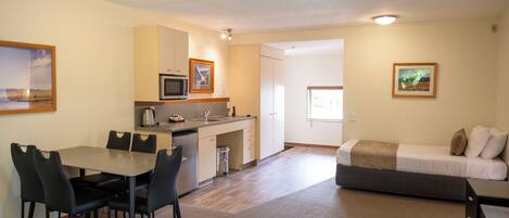 A compact hotel room with a kitchenette, a dining area with a table and chairs, a bed with pillows, and a small desk.